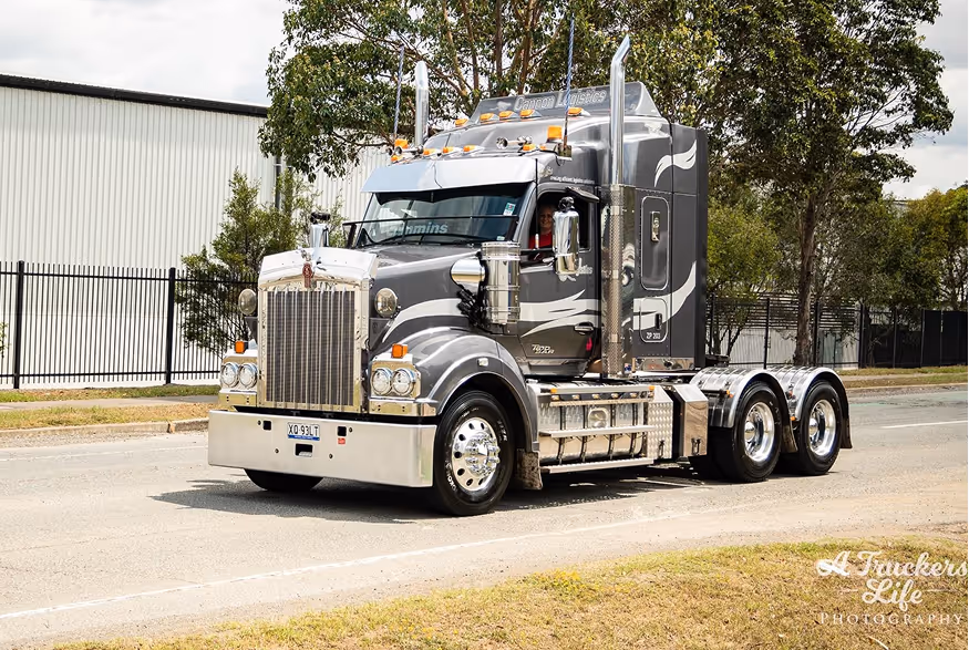 Gray Kenworth semi-truck cab with chrome details parked on a street near a fence and trees.