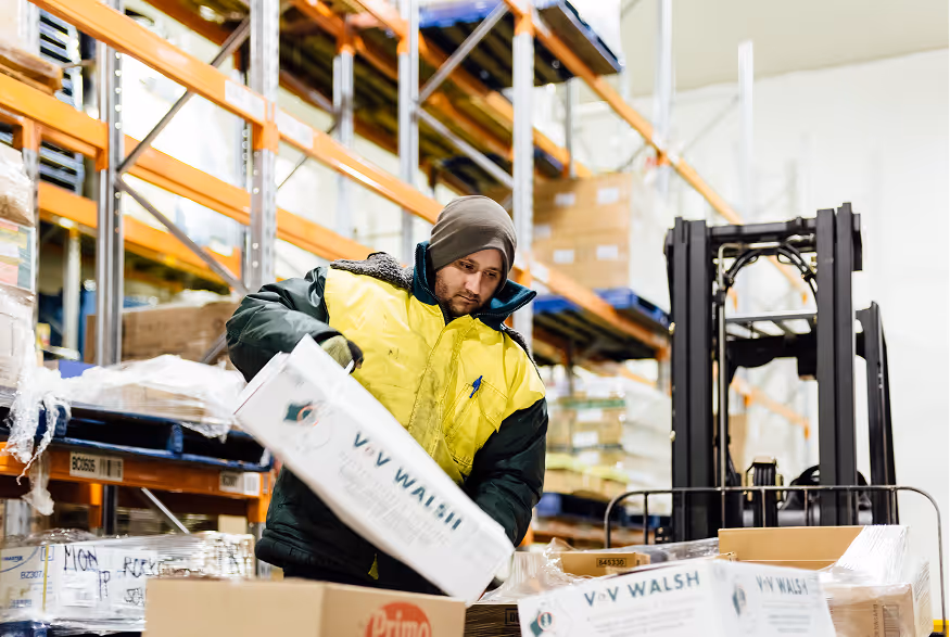 Warehouse worker in yellow jacket moving a box among stacked pallets and forklift in background.