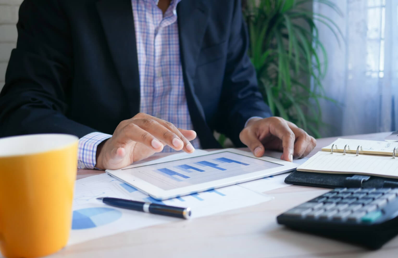 Person in business attire reviewing bar charts on a tablet, surrounded by documents, pen, calculator, and a yellow cup on a desk.