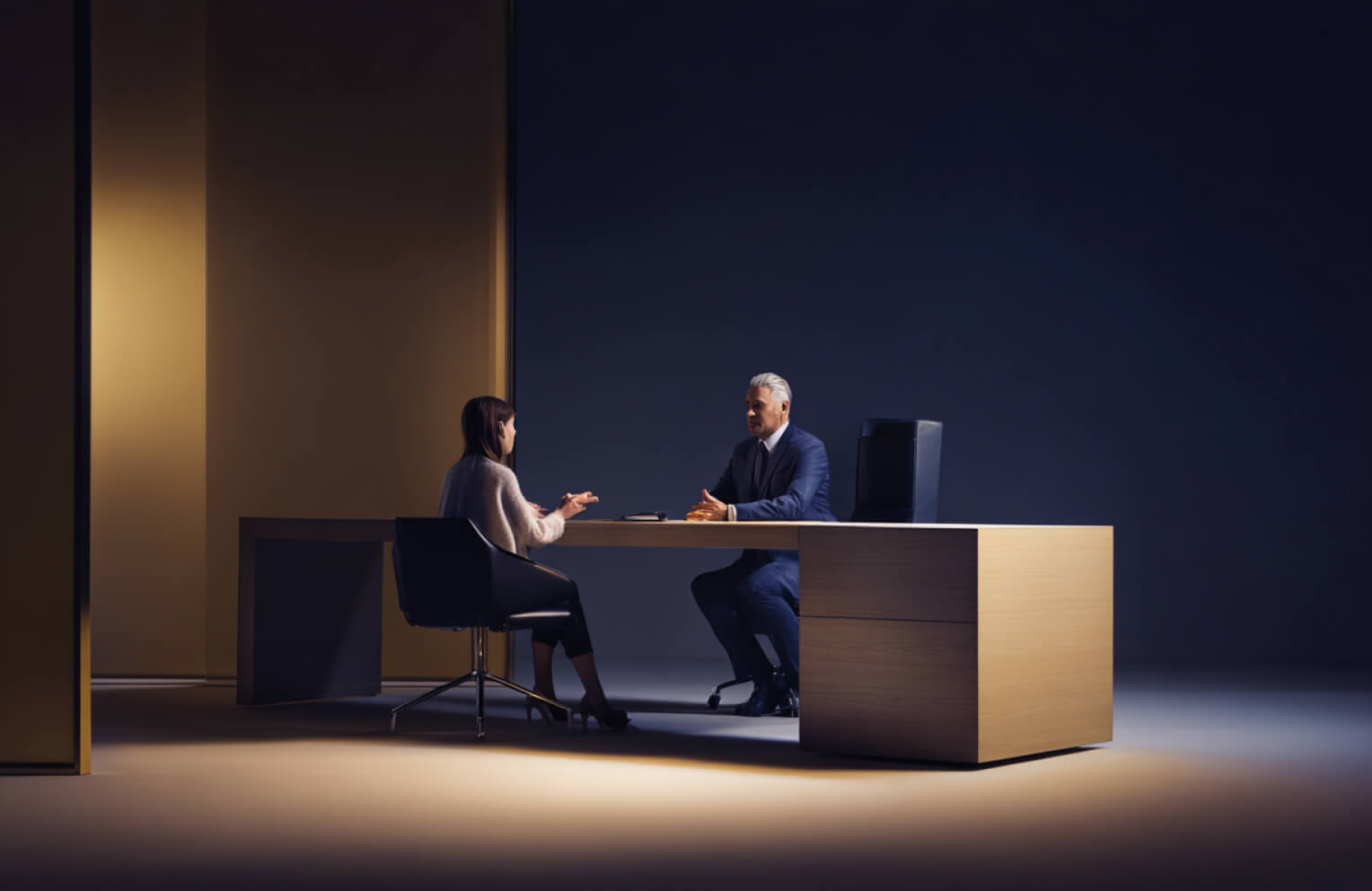 A professional business meeting between a man in a suit and a woman seated across a wooden desk in a dimly lit office.