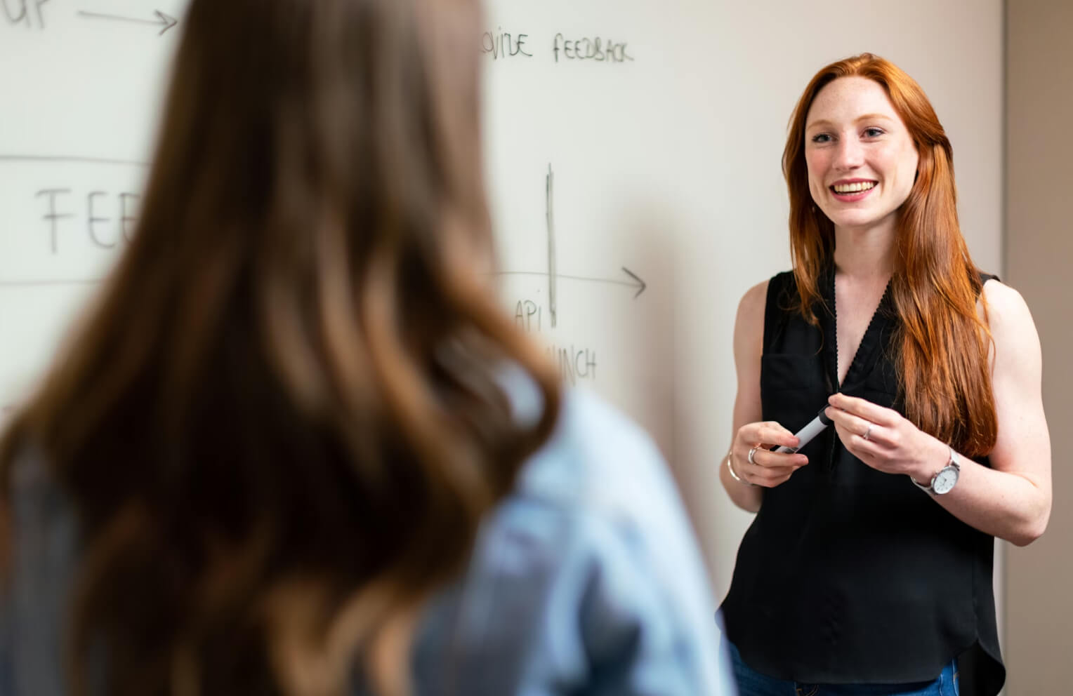 Smiling woman with red hair holding marker and talking to another person in front of whiteboard with diagrams and text.