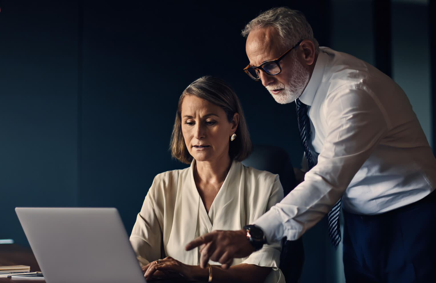 Mature businessman in glasses pointing at a laptop screen while advising a seated female colleague in an office.