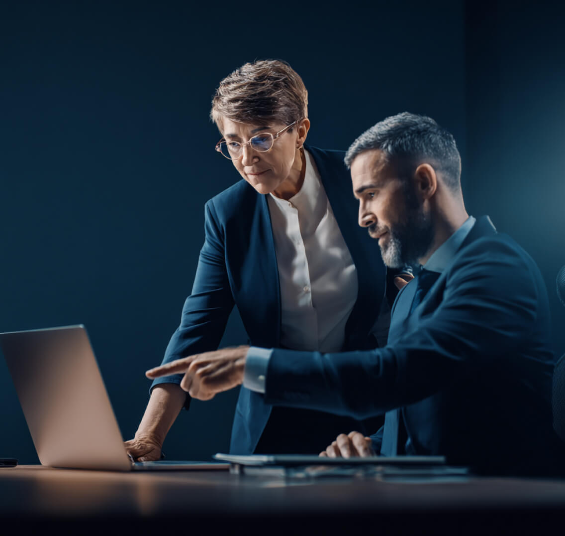 Two business professionals, a man and a woman, discussing work while looking at a laptop screen in a dimly lit office.