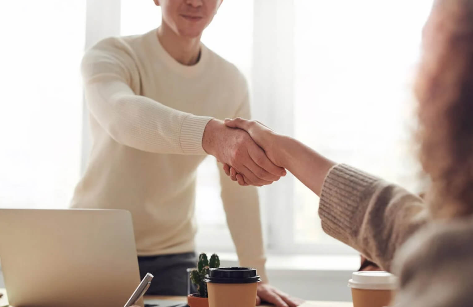 Two people shaking hands over a desk with a laptop and coffee cups, indicating agreement or negotiation.