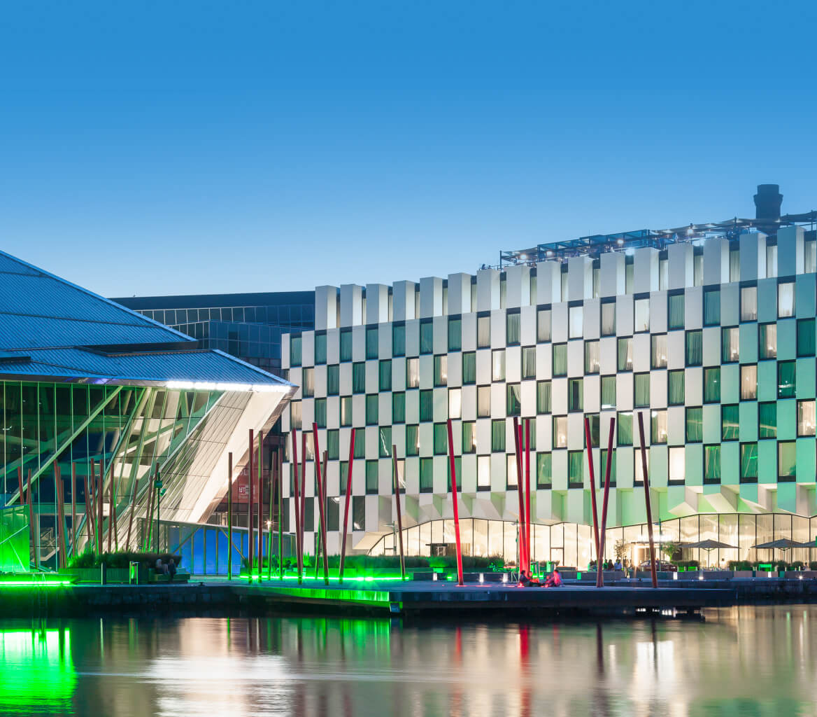Modern waterfront buildings with illuminated windows and green lights reflecting on the calm water at dusk.