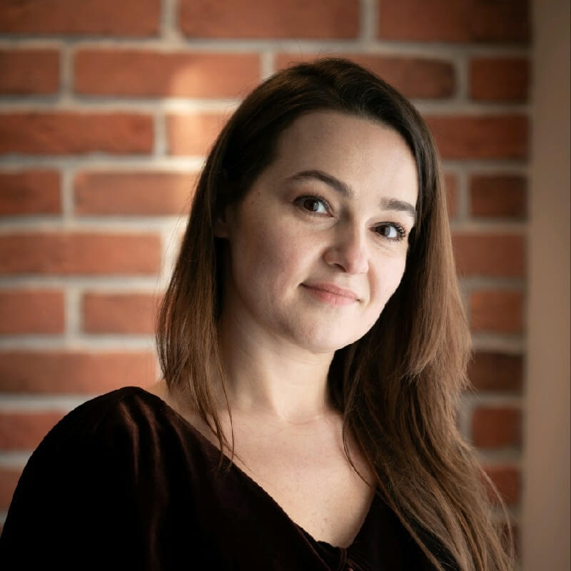 Portrait of a woman with long brown hair wearing a dark top, standing in front of a brick wall.