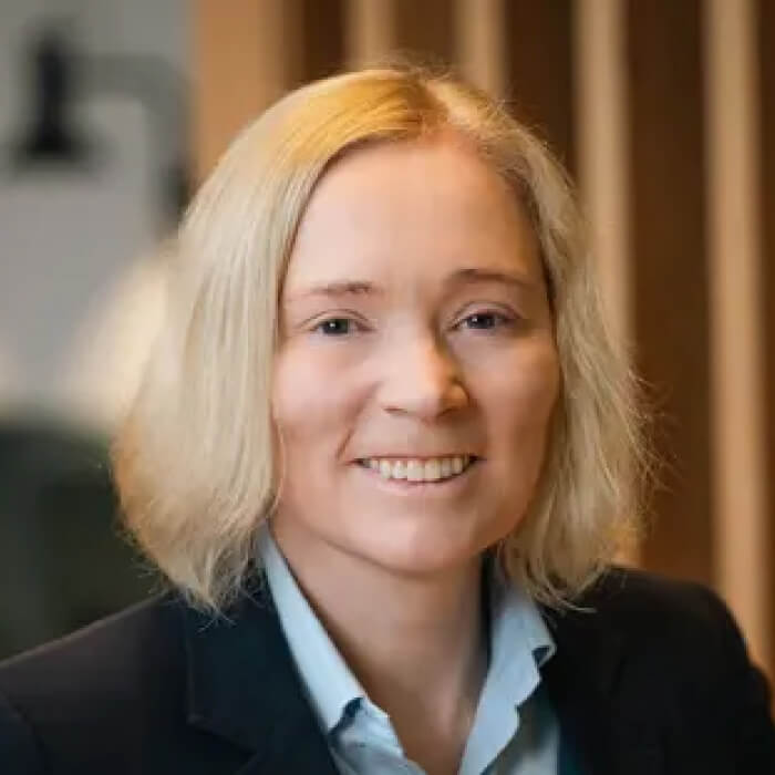 Smiling woman with shoulder-length blonde hair wearing a light blue collared shirt and dark blazer.