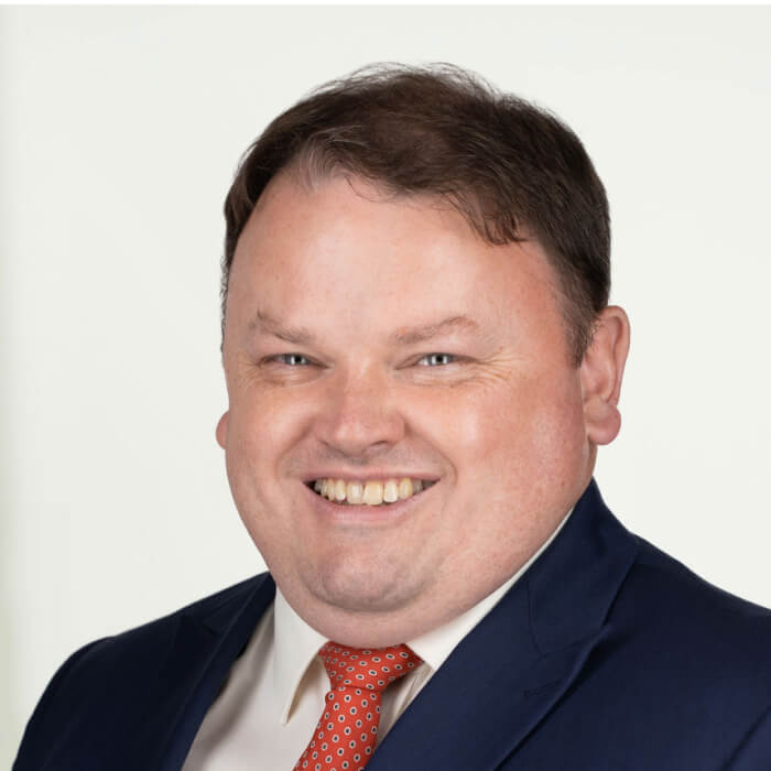 Smiling man wearing a dark blue suit, white shirt, and red patterned tie against a plain background.