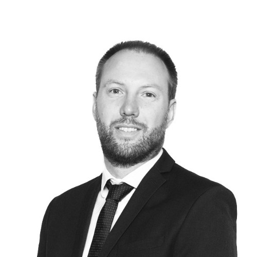 Man with short hair and beard wearing a dark suit, white shirt, and patterned tie against a white background.