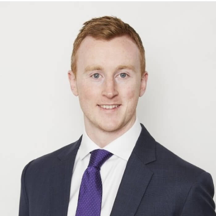Smiling man with short red hair wearing a dark suit, white shirt, and purple tie against a plain light background.