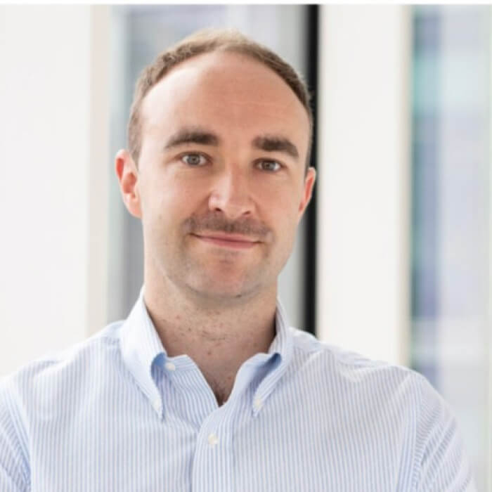 Portrait of a man with short light brown hair and a slight smile, wearing a light blue striped button-up shirt.