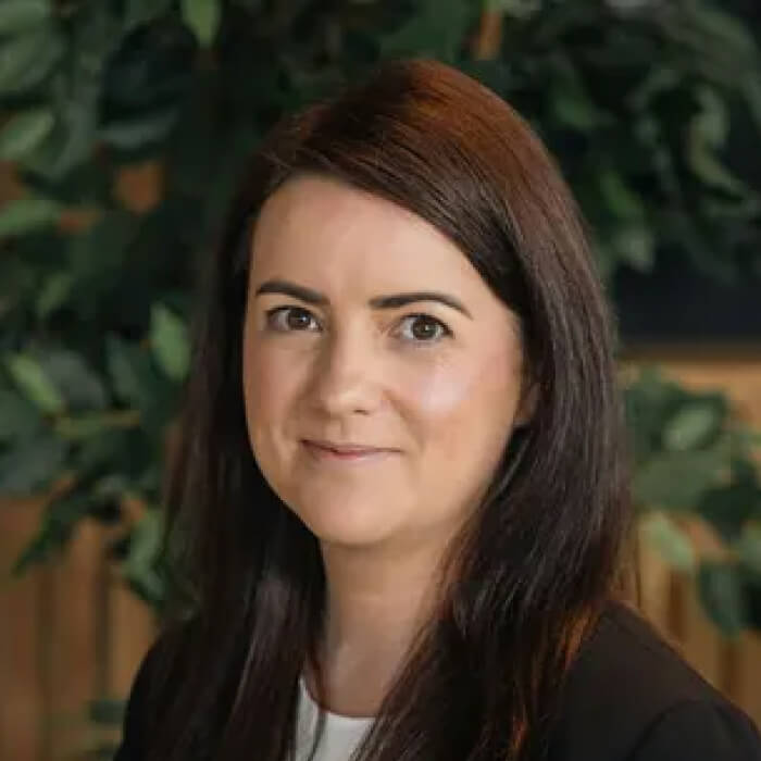 Portrait of a woman with long dark hair wearing a black blazer and white top, with a blurred green and brown background.