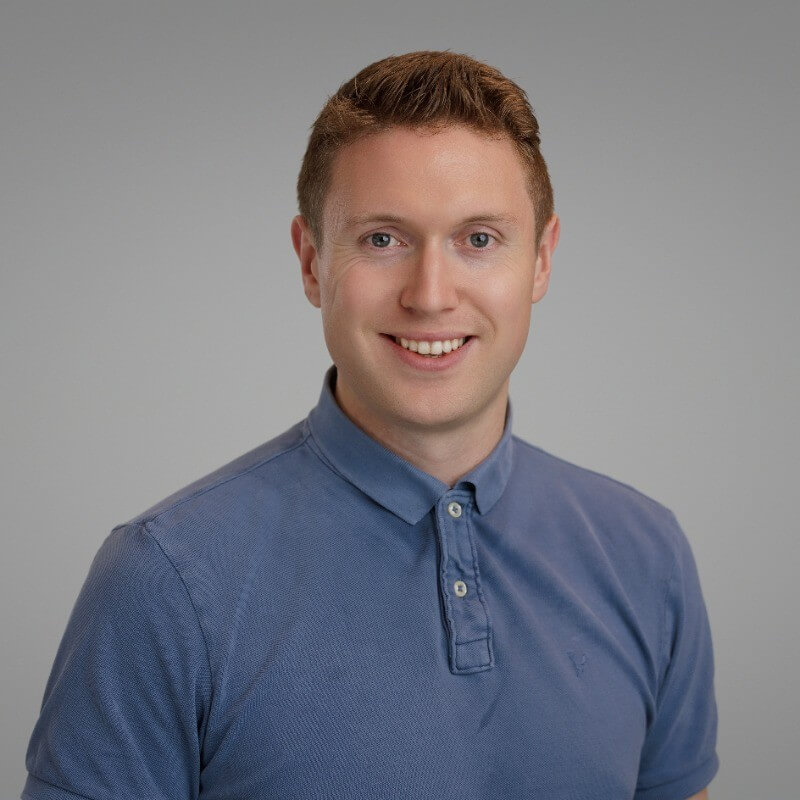 Smiling young man with short brown hair wearing a blue collared shirt against a plain gray background.