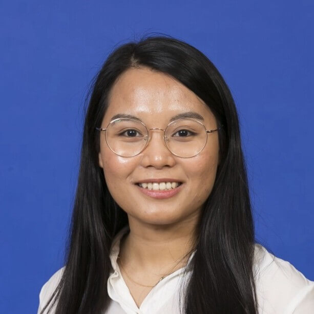 Smiling woman with long black hair, wearing round glasses and a white shirt, against a blue background.