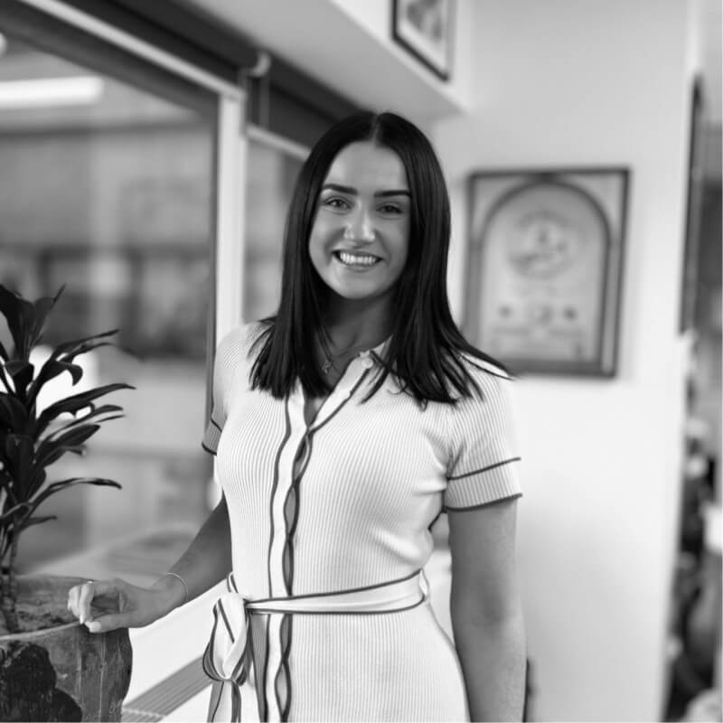 Smiling woman with straight dark hair wearing a belted, short-sleeve dress standing inside near a window and plant.