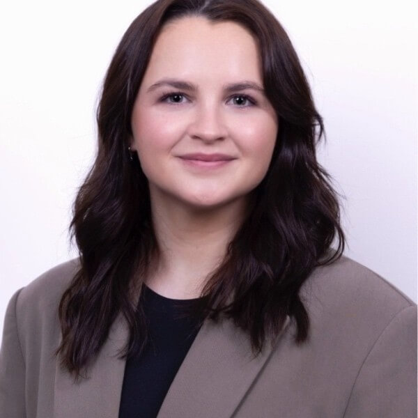 Portrait of a woman with long dark hair wearing a taupe blazer and black top, smiling softly against a white background.
