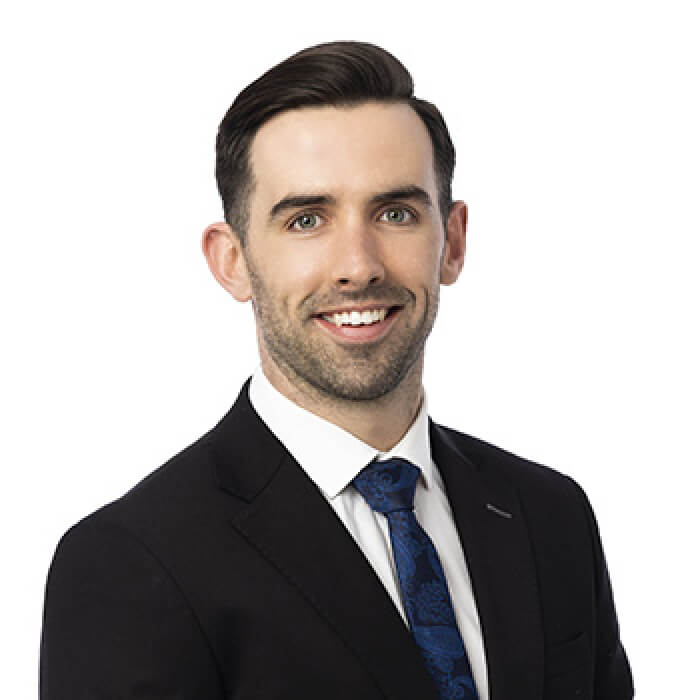 Smiling man with short dark hair wearing a black suit, white shirt, and blue patterned tie against a white background.