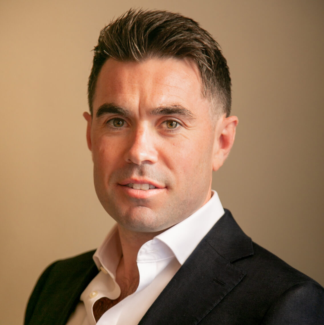 Headshot of a man with short dark hair, wearing a white shirt and dark blazer, against a plain beige background.