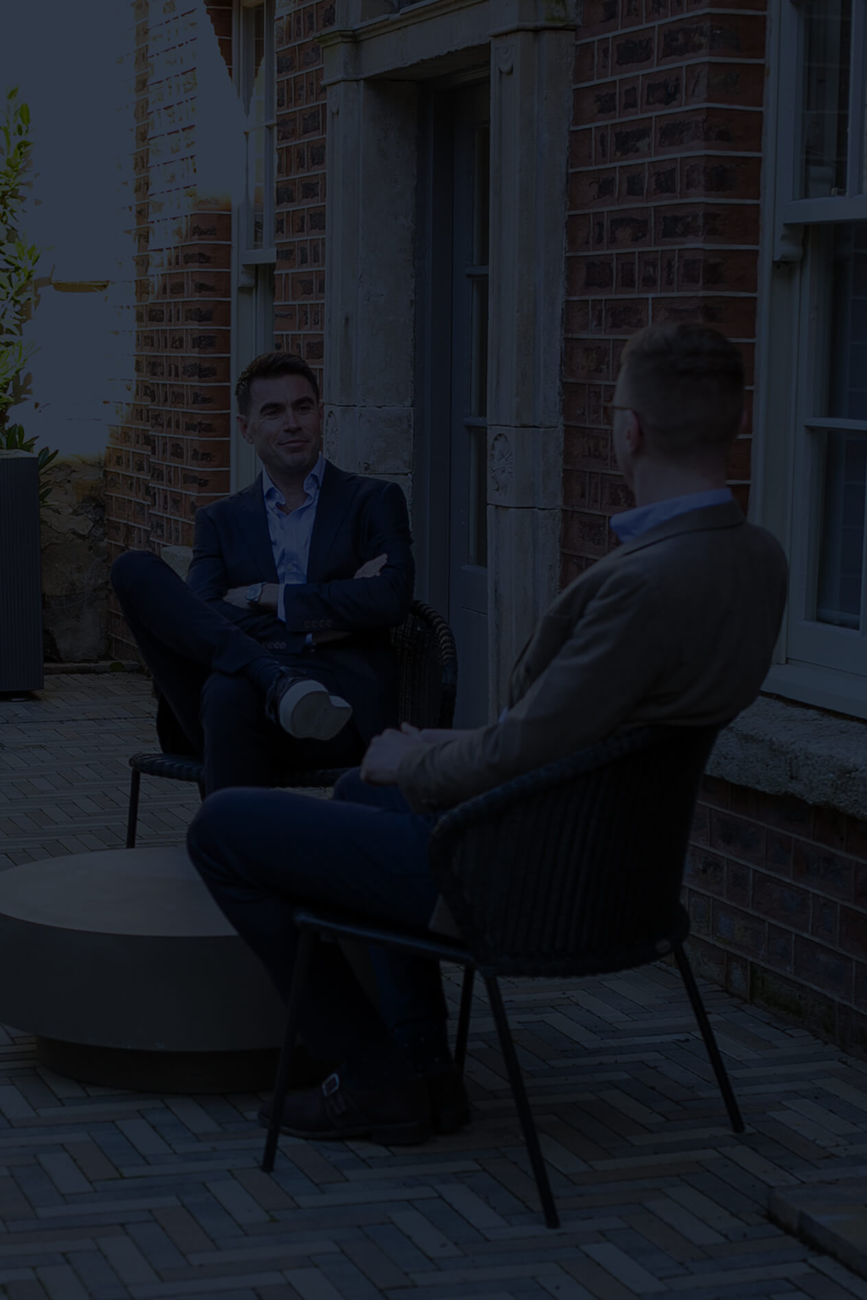 Two men in business attire sitting on wicker chairs having a conversation outside a brick building.