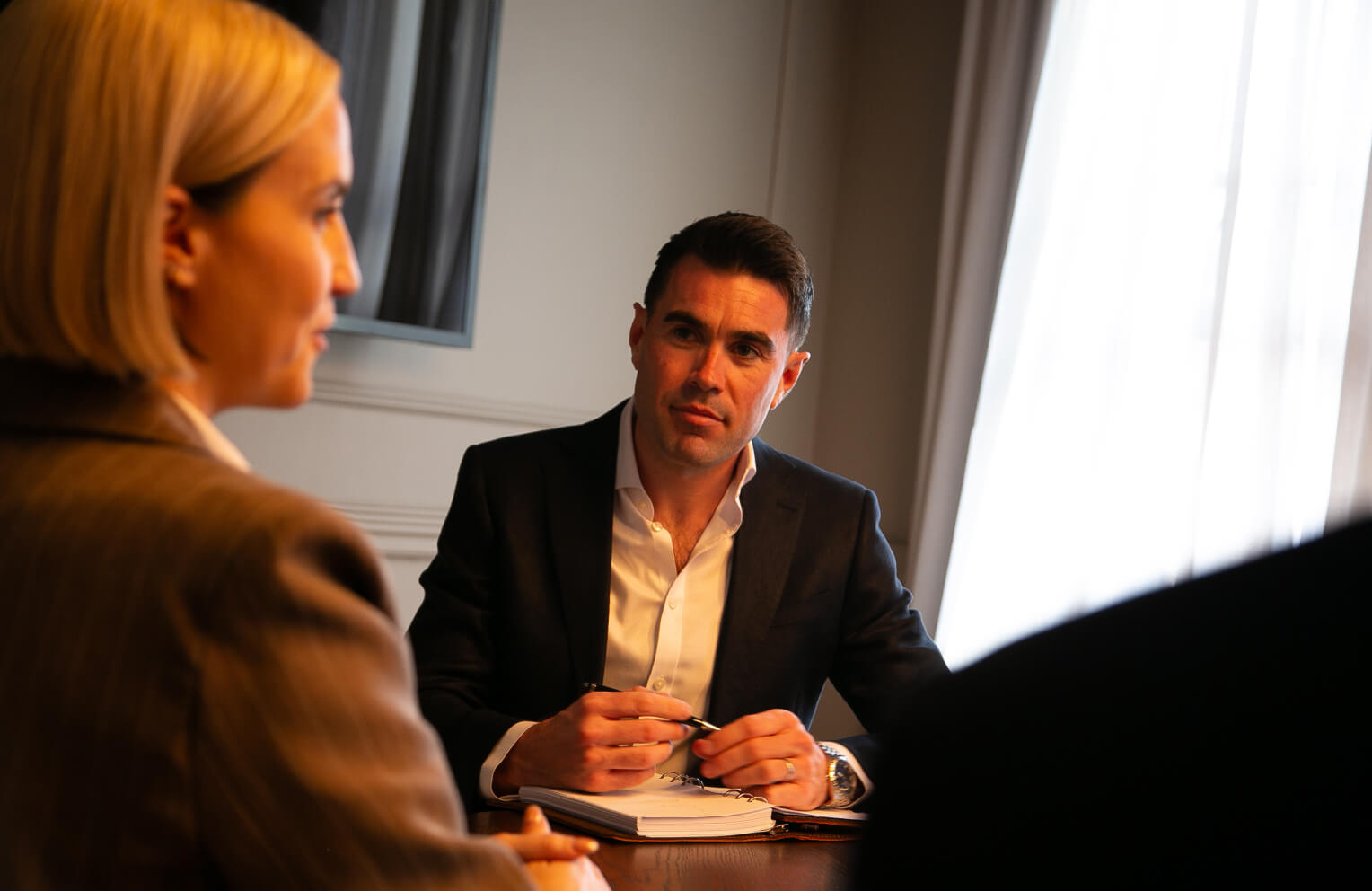 Man in a dark suit holding a pen and listening attentively to a woman in a brown blazer during a meeting.