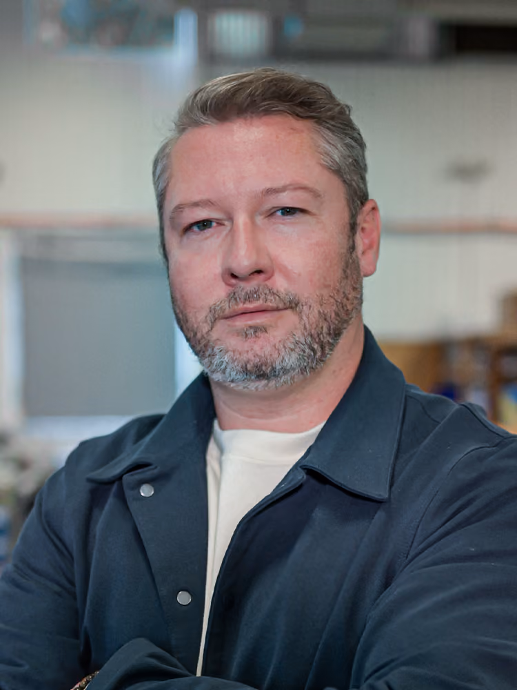 Man with short gray hair and beard wearing a dark jacket and white shirt indoors.