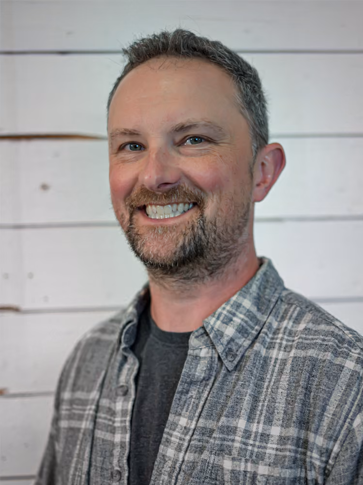 Smiling man with short gray hair and beard wearing a plaid shirt over a dark t-shirt.