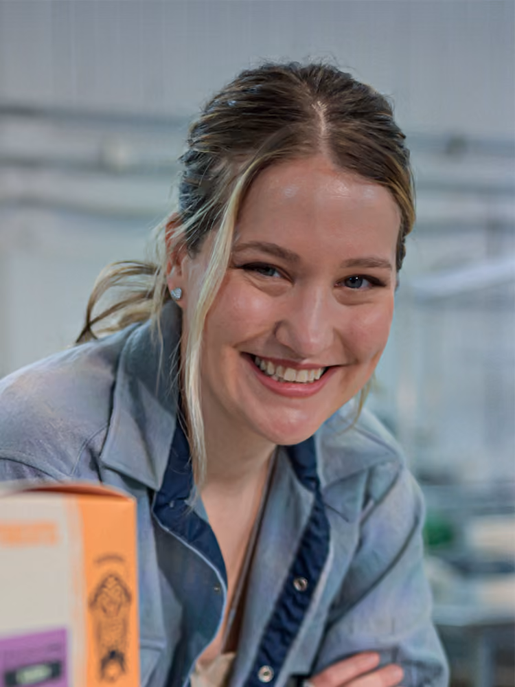 Smiling woman with blonde hair wearing a blue shirt leaning on a table in a workspace.