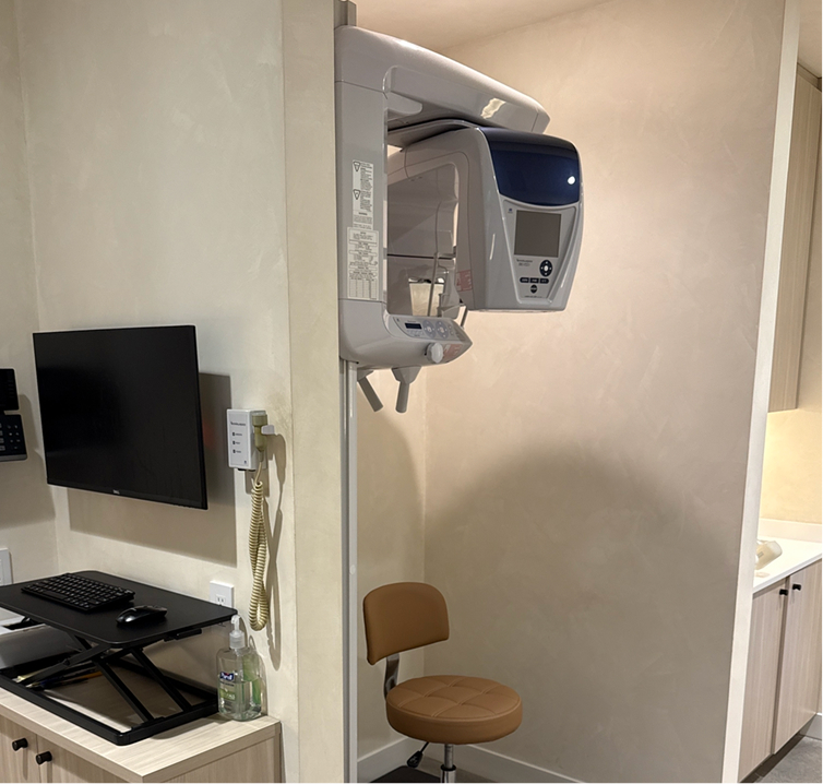Dental CT scan machine mounted on wall with brown stool and workstation with monitor and keyboard.