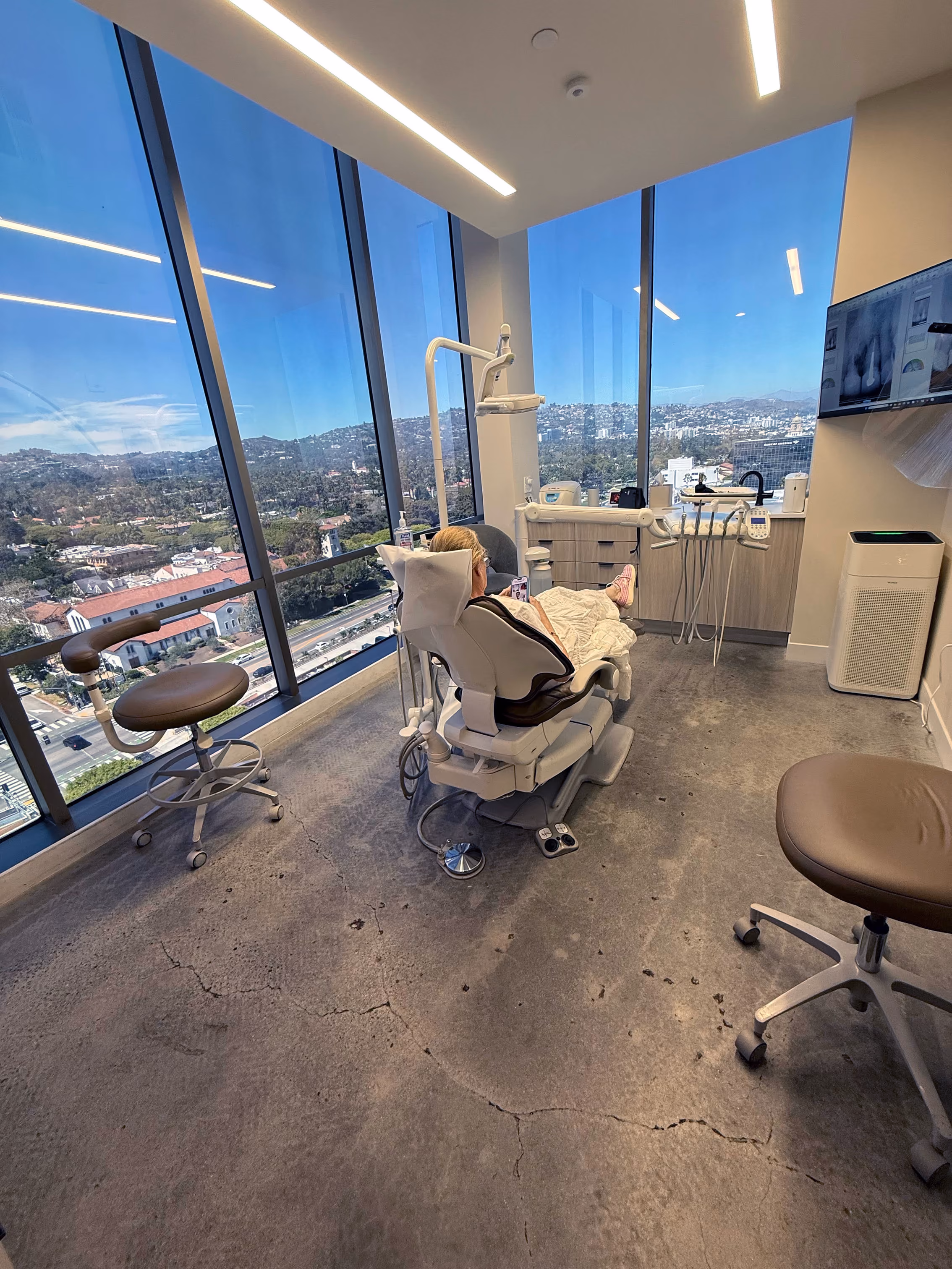 Patient sitting in a dental chair inside a modern clinic with large windows showing a cityscape outside.