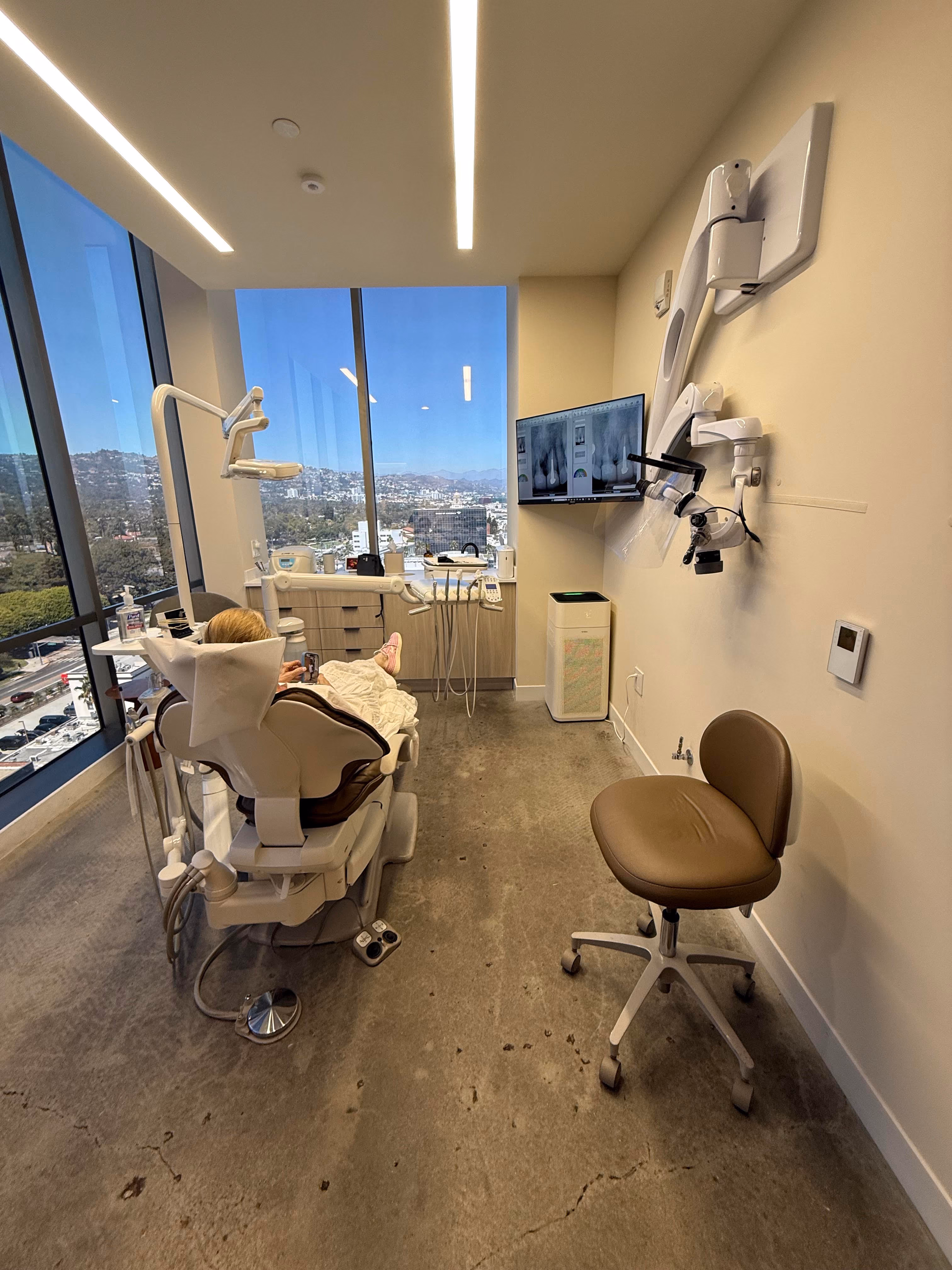 Modern dental office with patient in chair, dental equipment, and city view from large windows.
