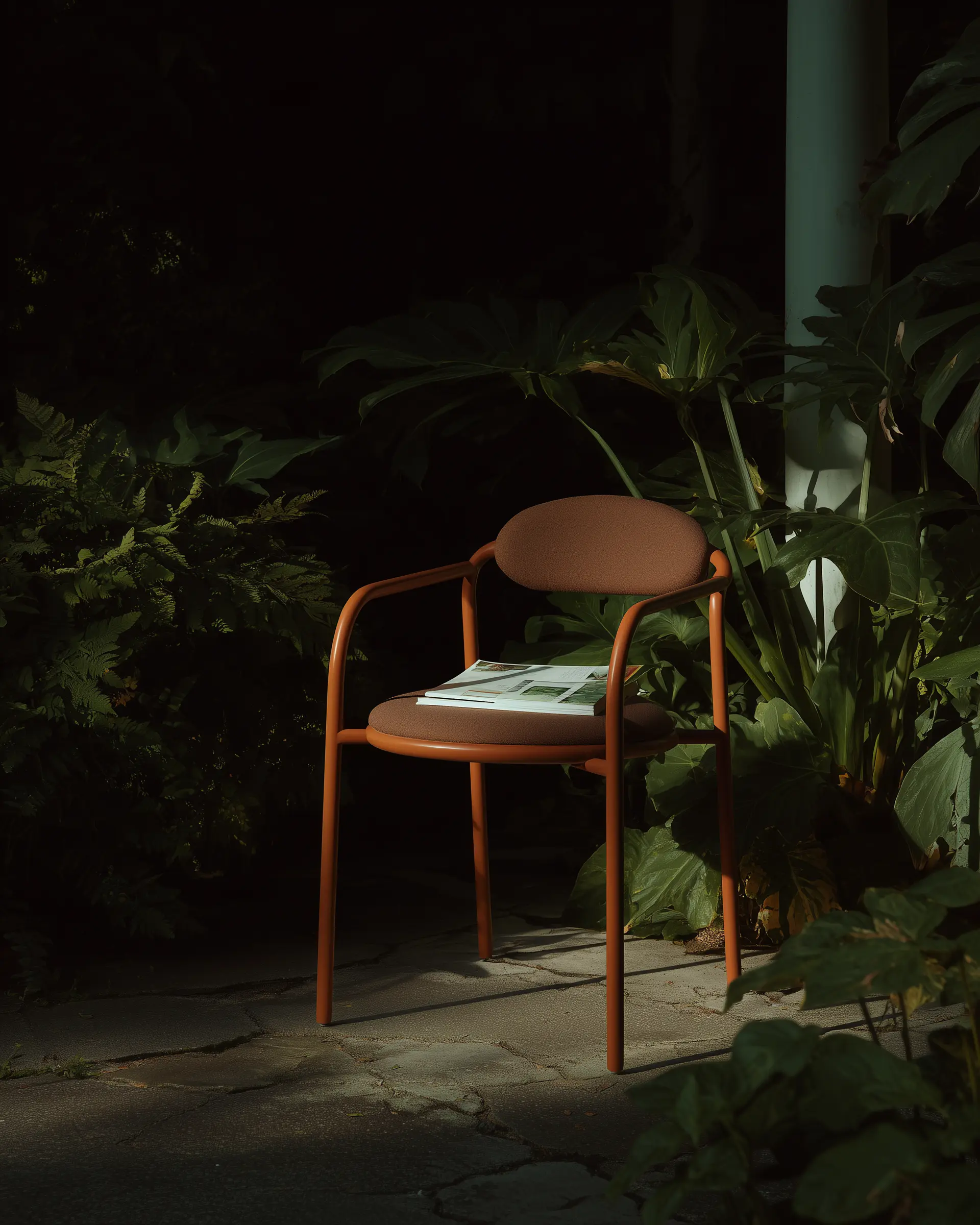 Brown cushioned chair with a reddish frame and an open magazine on the seat, placed outdoors on cracked pavement surrounded by large green leaves.