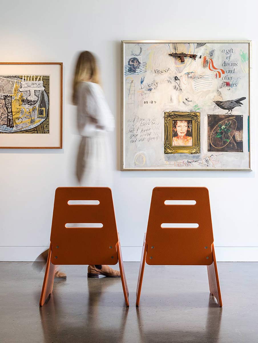 OA Brand image. Modern gallery setting with two orange chairs facing a white wall adorned with framed abstract and mixed media artworks, and a blurred figure walking by.