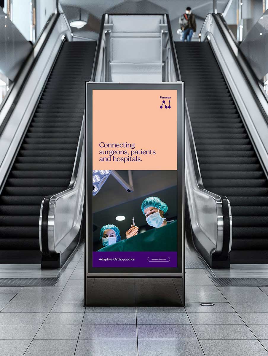 Panacea Ai Brand image. A Digital display between escalators showing an ad with surgeons in masks and caps, text reading 'Connecting surgeons, patients and hospitals.'