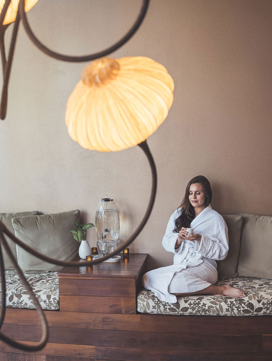 Gwinganna Brand image. Woman in a white bathrobe sitting on a floral cushioned bench holding a cup, with a water dispenser and small candles on a wooden table beside her.