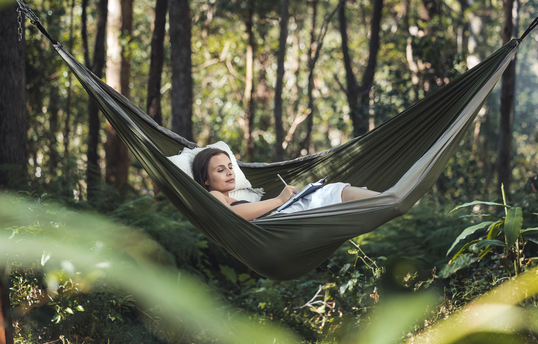 Gwinganna Brand image. Woman writing in a notebook while reclining on a pillow in a hammock in a forest.