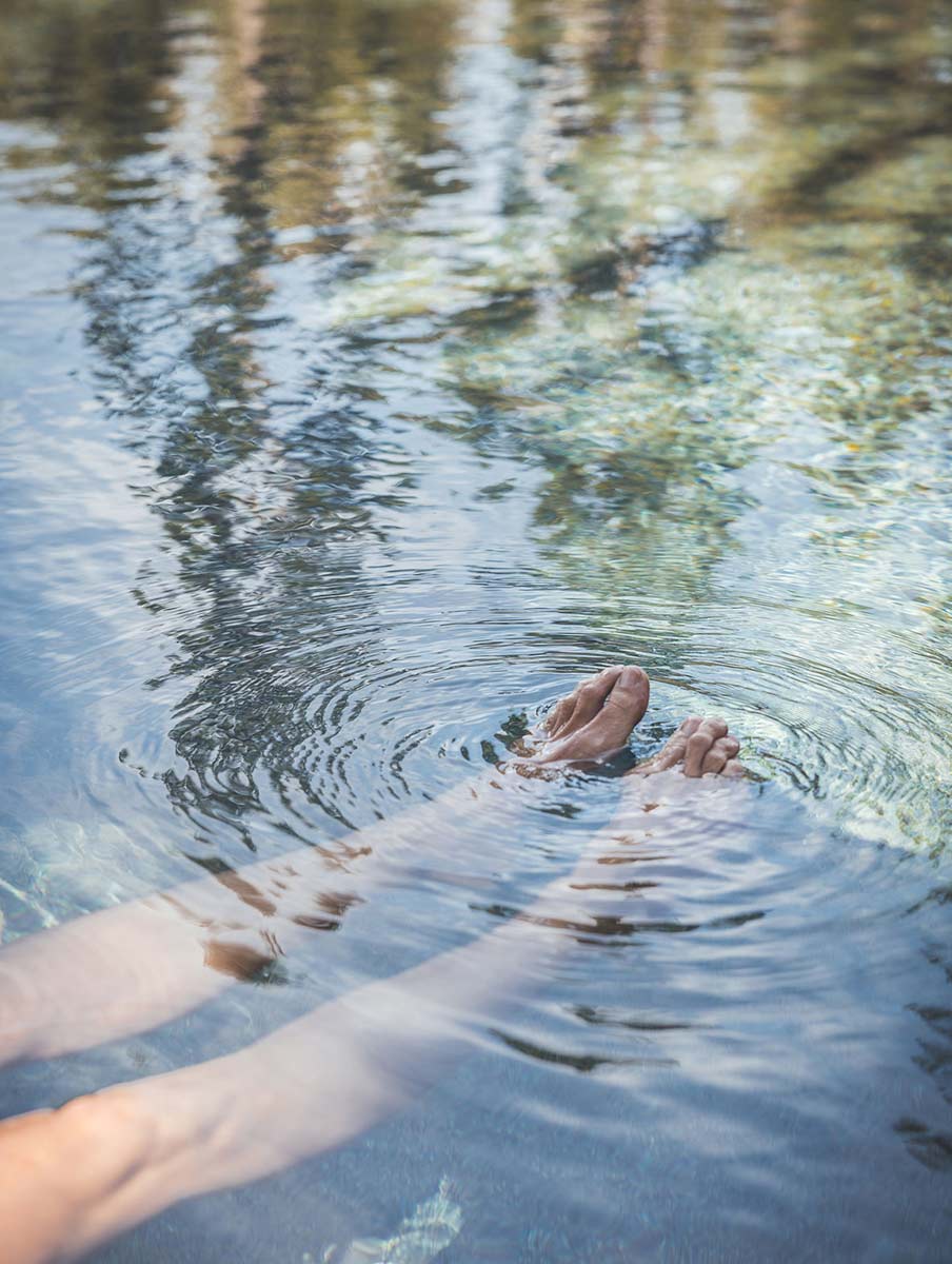 Gwinganna Brand image. Person's legs and feet submerged in clear water with ripples and reflections of trees on the surface.