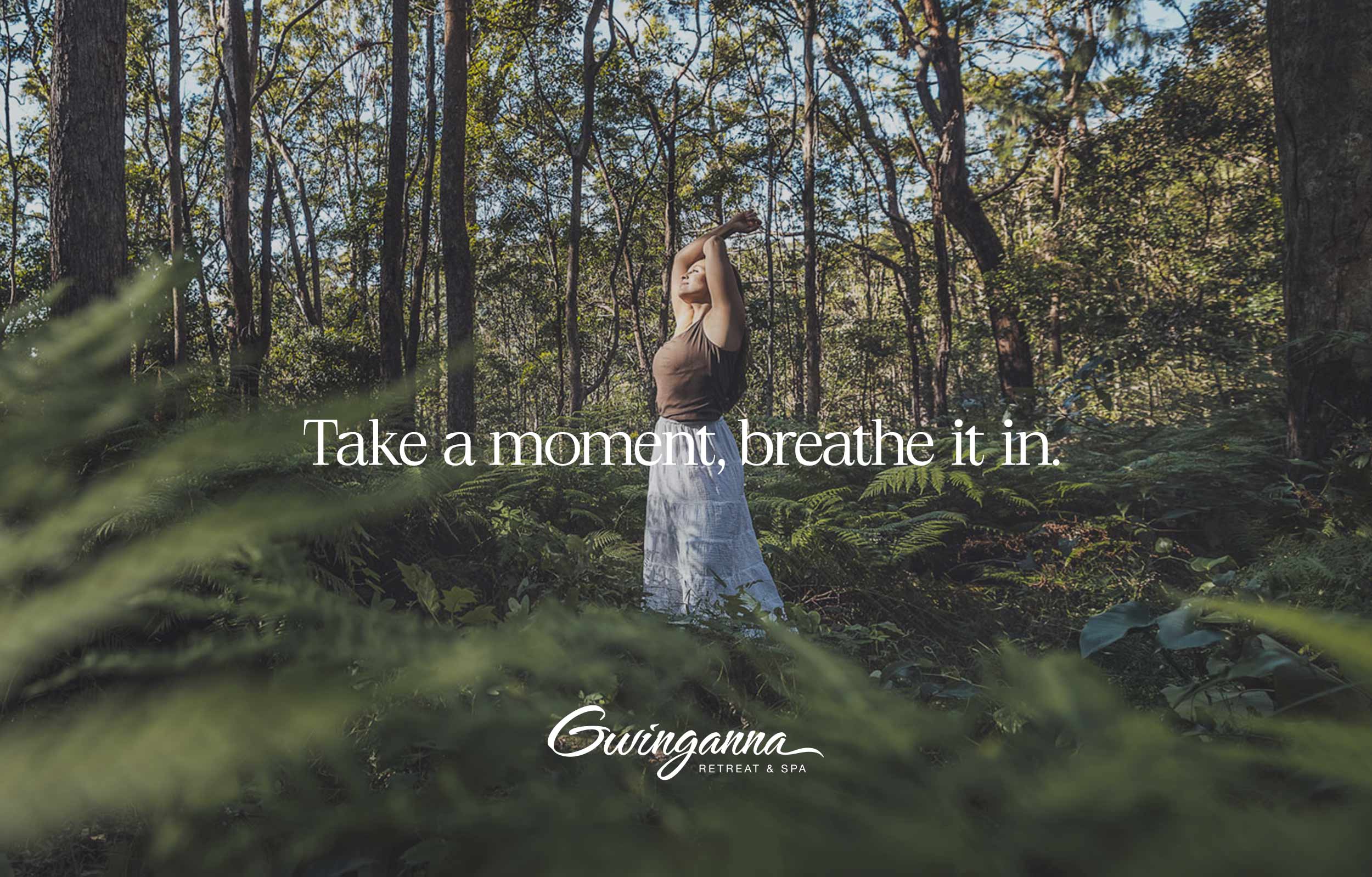 Gwinganna Brand image. Woman standing in a lush forest with arms raised, surrounded by greenery and trees.