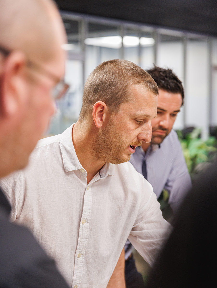 IEDM Team. Three men in a discussion in an office setting, with one man in a white shirt speaking and two others attentively listening.