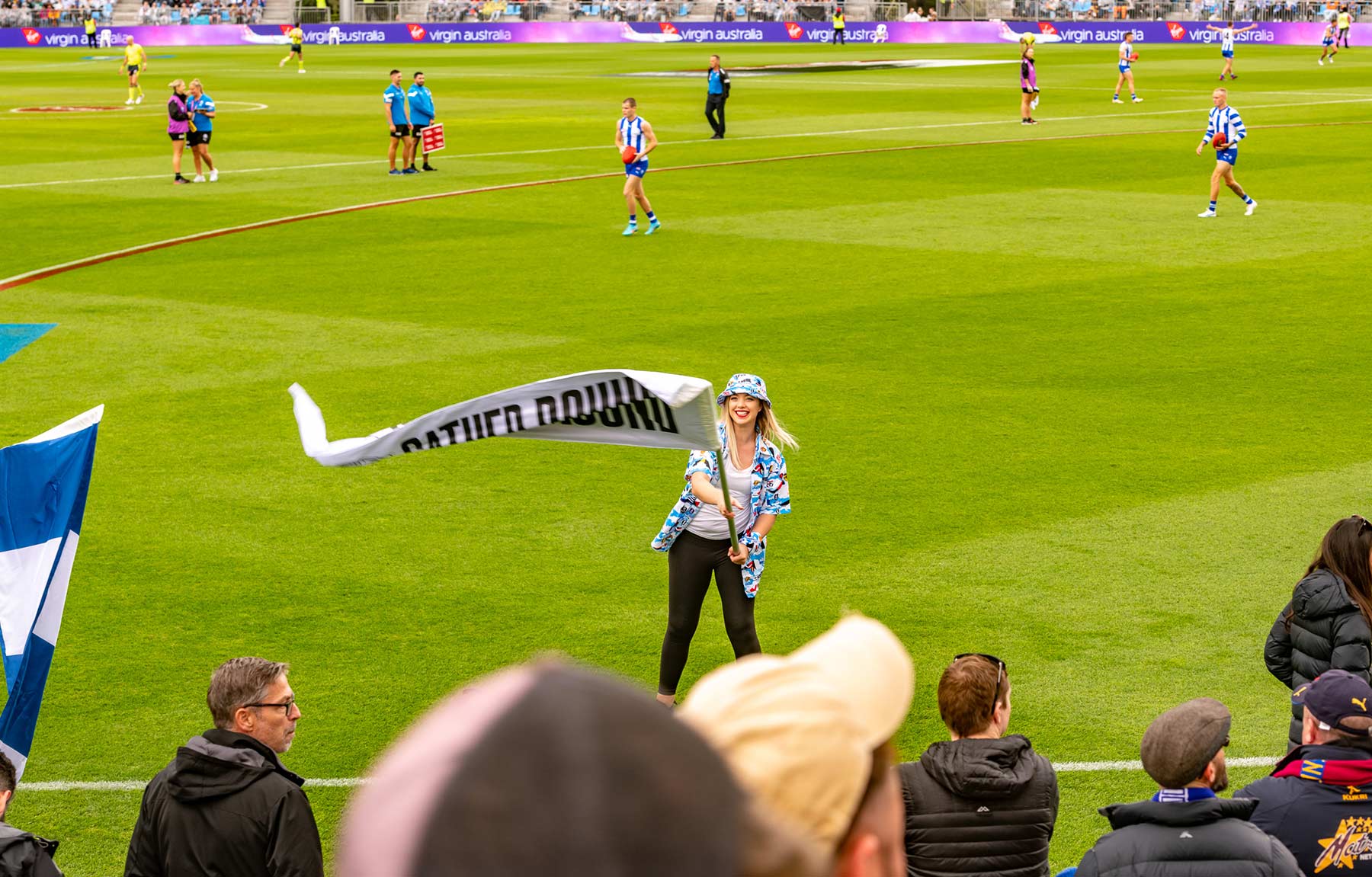 IEDM Gather Round. Woman waving a flag on a football field with players and spectators in the background.