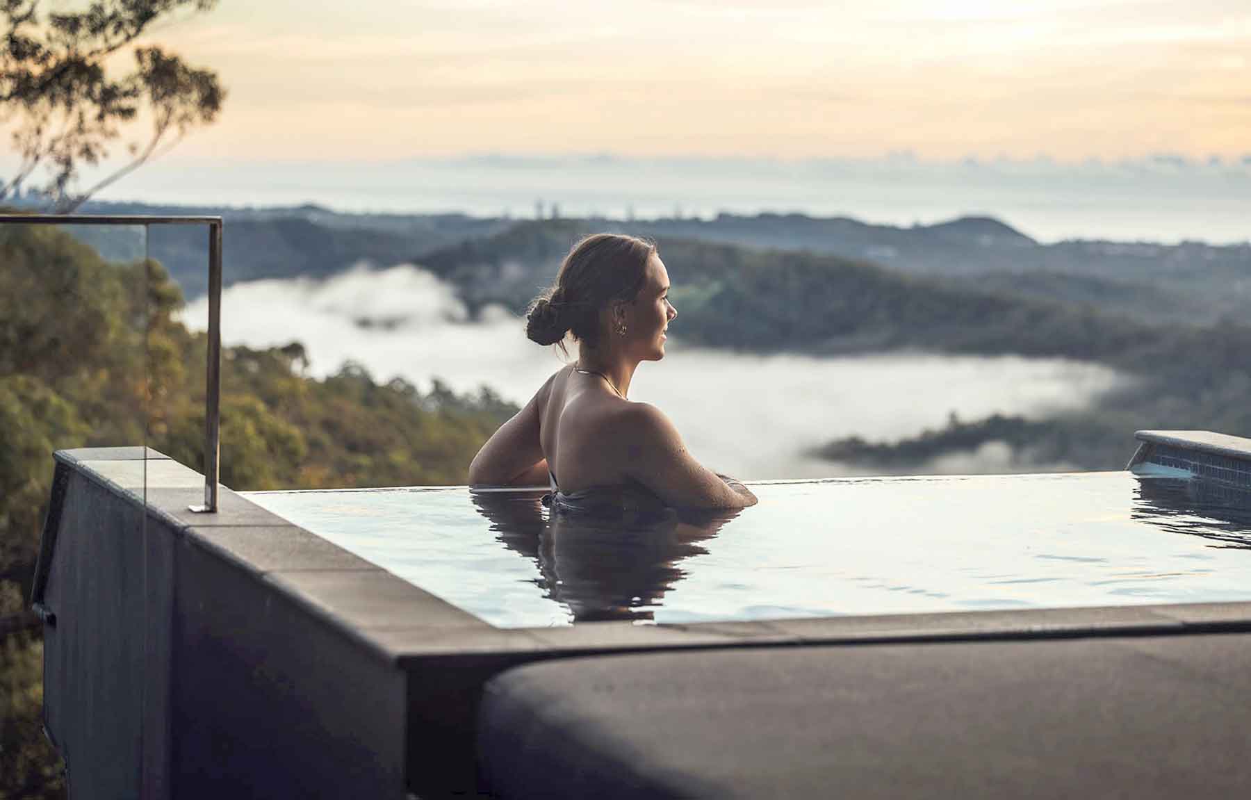 Gwinganna brand image. A woman relaxing in an infinity pool overlooking misty hills and forest at sunrise or sunset.