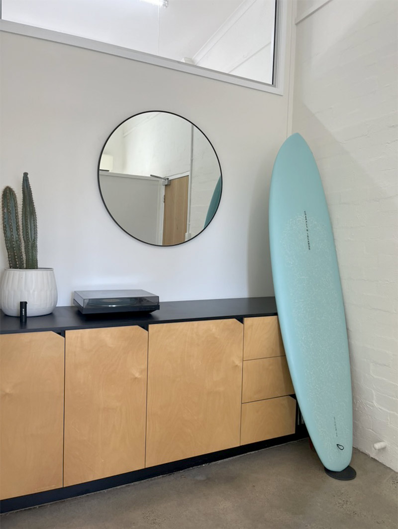 Mariachi Creative Studio Gold Coast. Minimalist interior with wooden cabinets, a round wall mirror, a cactus plant in a white pot, a record player, and a blue surfboard leaning against a white brick wall.