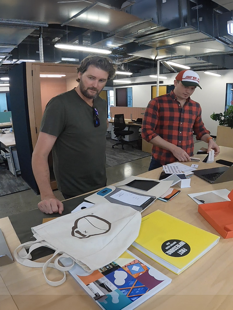 Two men standing at a desk in a modern office, examining papers and a laptop with notebooks and a tote bag on the desk.