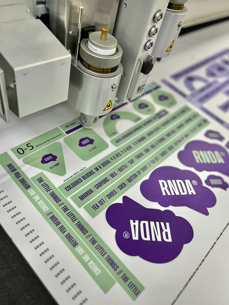 Close-up of a printing machine applying purple and green ink on paper with text and shapes related to the brand RNDA.