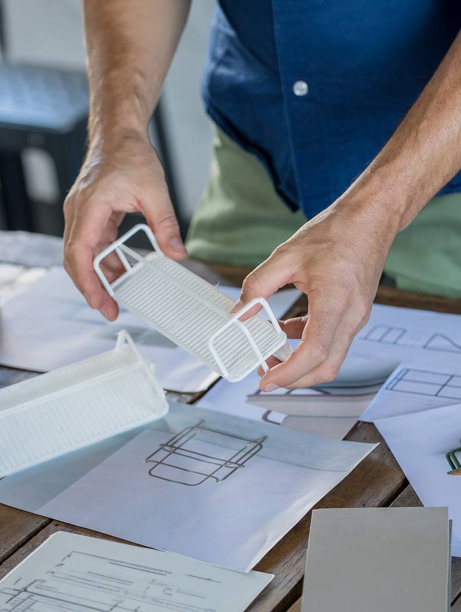 Outside Assembly. A person holding a white 3D-printed model over technical drawings on a wooden table.