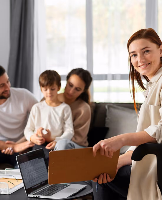 Female professional smiling while holding a clipboard in a living room with a couple and child seated on a couch.