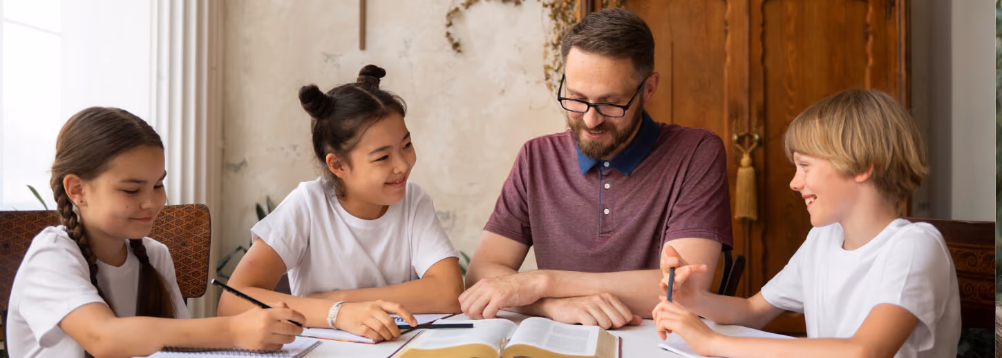 A man with glasses teaching or studying with three smiling children around a table with open books and notebooks.