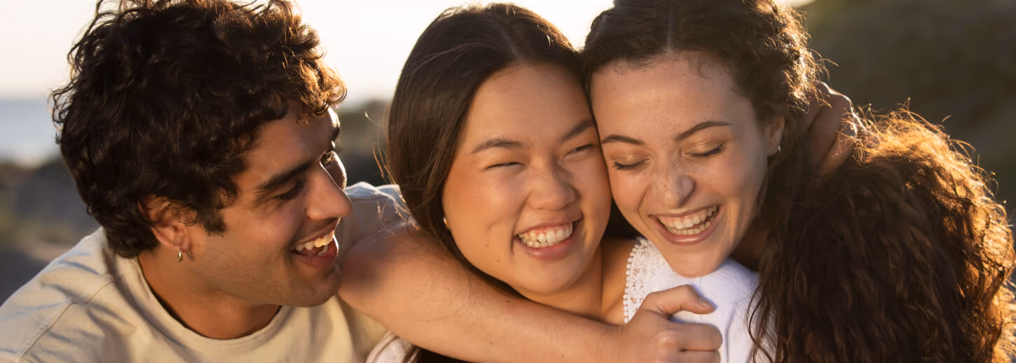 Three friends smiling and embracing each other outdoors in warm sunlight.