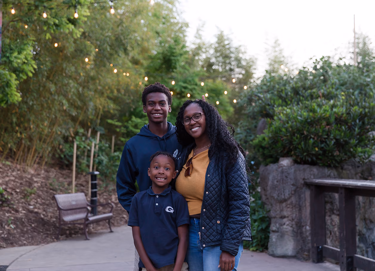 Smiling woman and two boys standing outdoors with string lights and greenery in background.