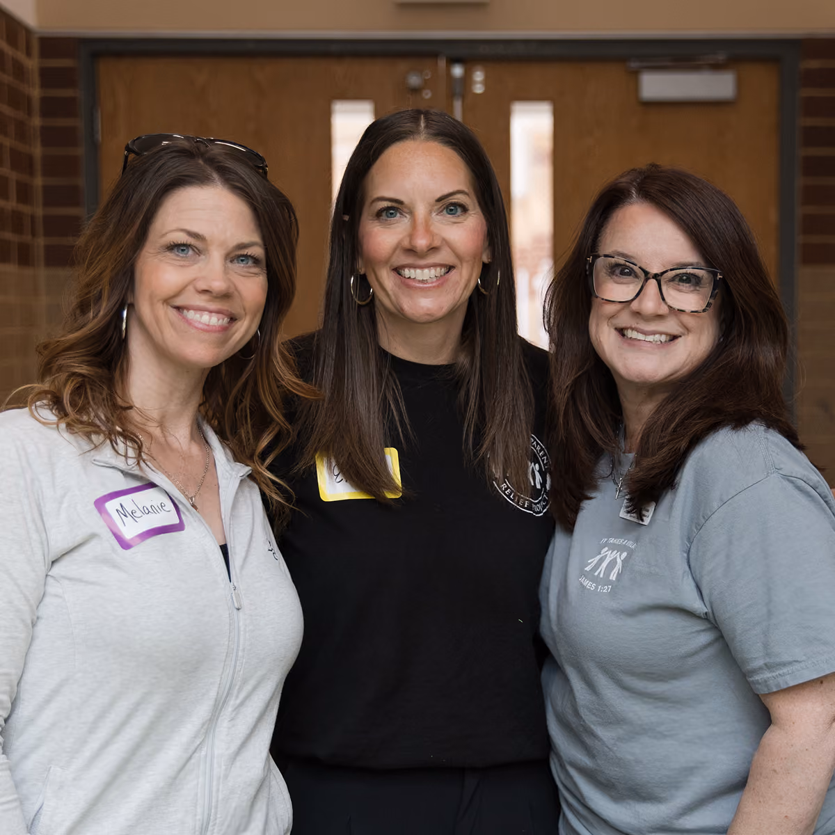 Three smiling women standing close together indoors, wearing casual tops and name tags.