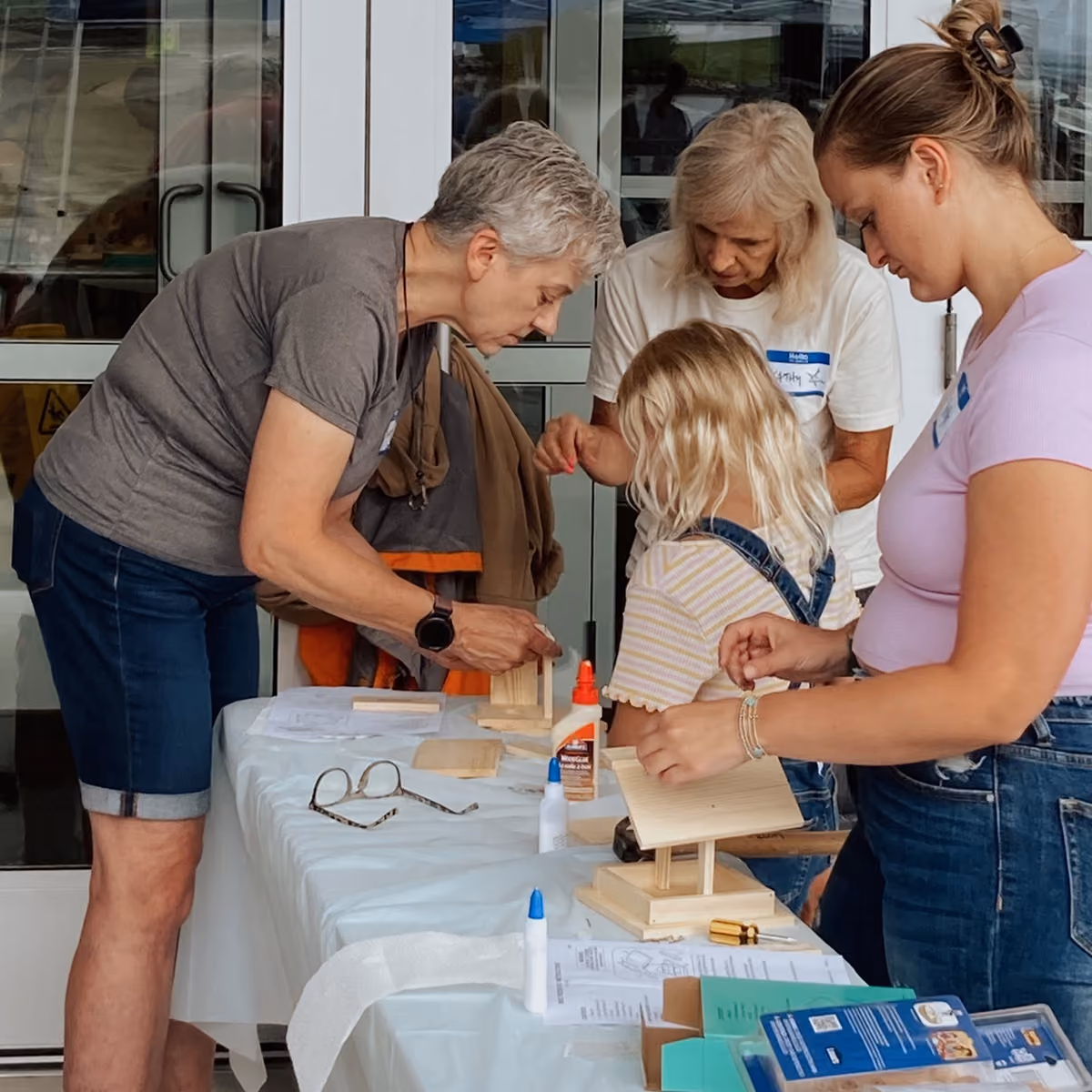 Three women and a girl engaged in assembling small wooden birdhouses at a table with glue and tools.